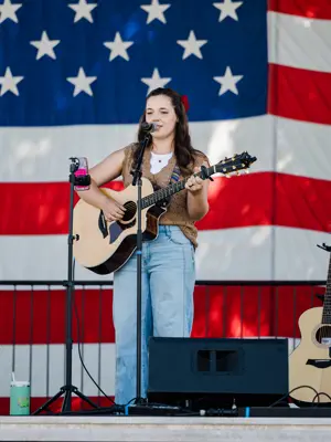 Young lady playing acousitc guitar on stage with big american flag in the background