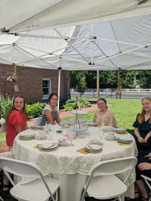 Youth sitting at a tea party table with green grass and orange brick whitaker home in the background on a sunny summer day